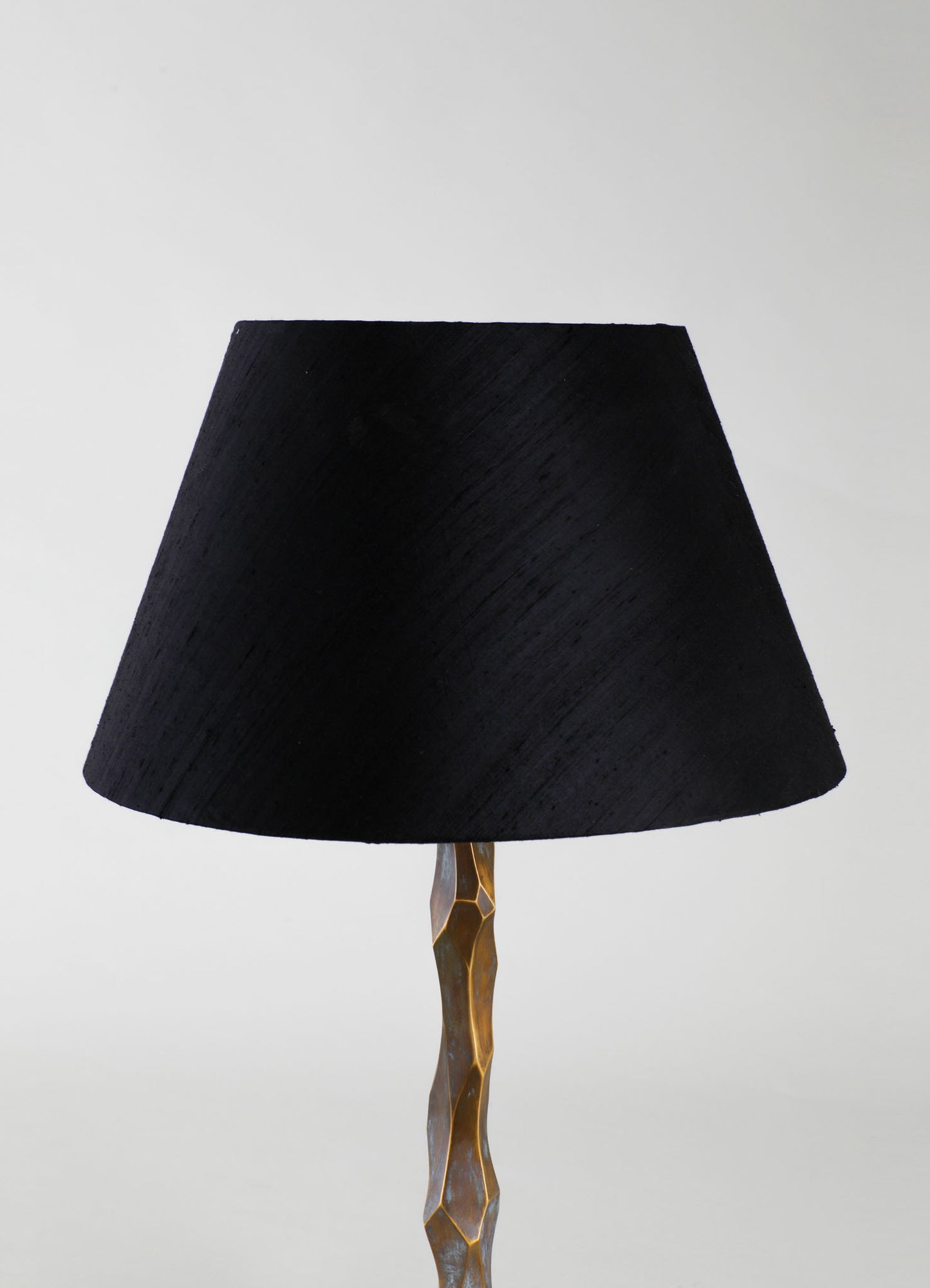 A table lamp with a black fabric lampshade featuring a textured, slightly conical shape, mounted on a sculptural, metallic gold base with irregular geometric facets.