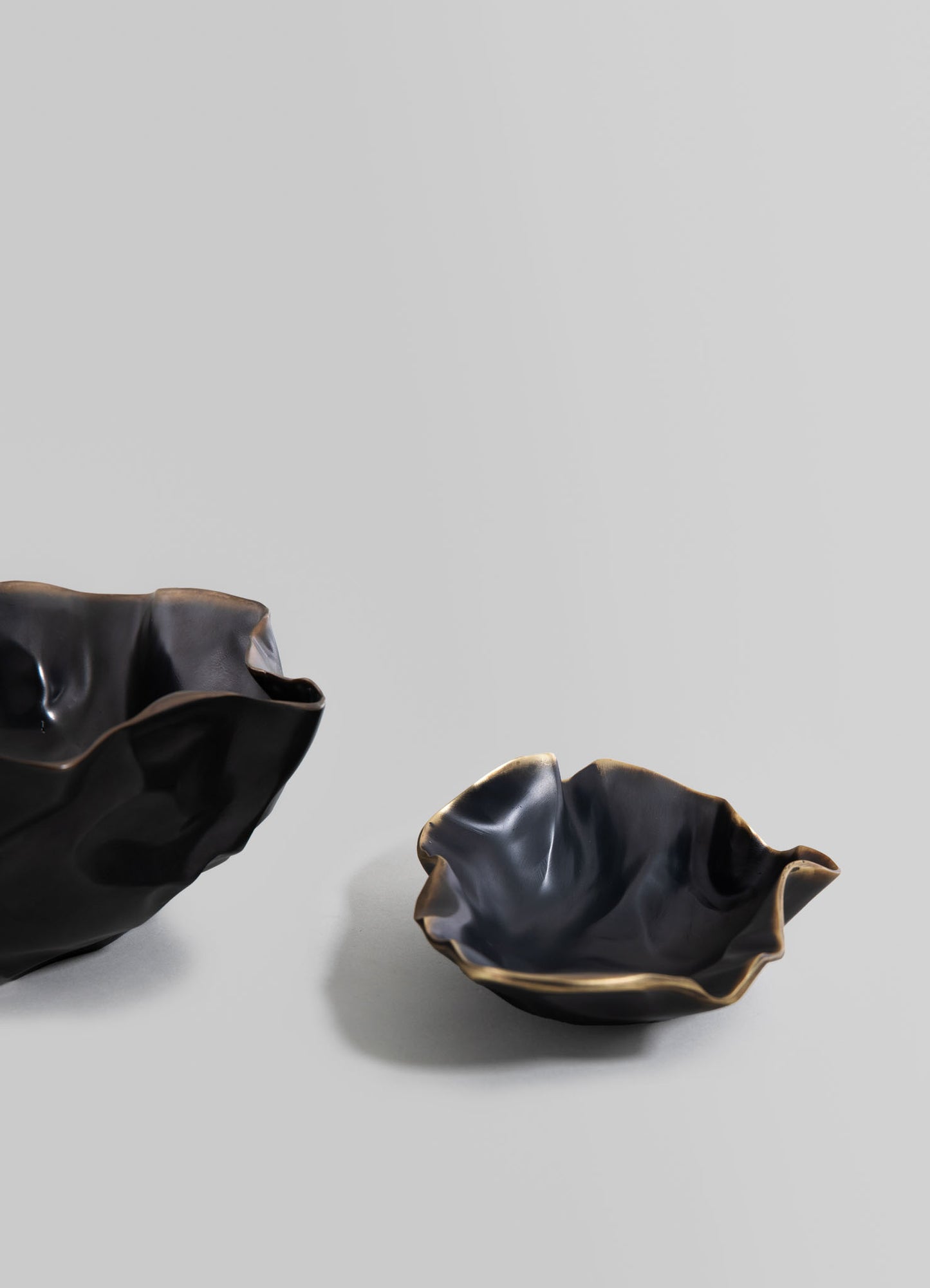 Two ceramic bowls with irregular, ruffled edges in glossy black and beige tones, featuring a fluid, organic form, set against a plain light gray background.