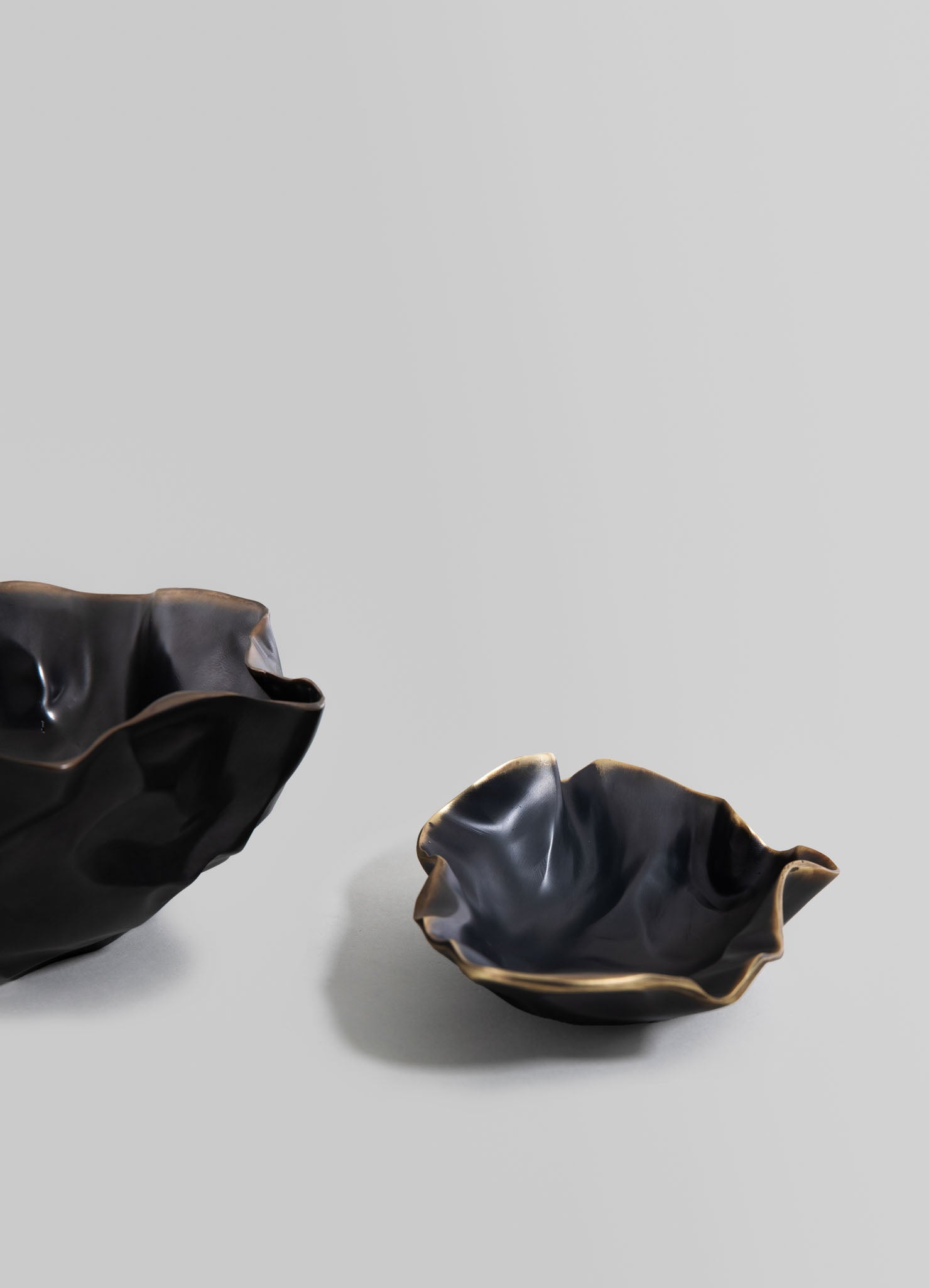 Two ceramic bowls with irregular, ruffled edges in glossy black and beige tones, featuring a fluid, organic form, set against a plain light gray background.