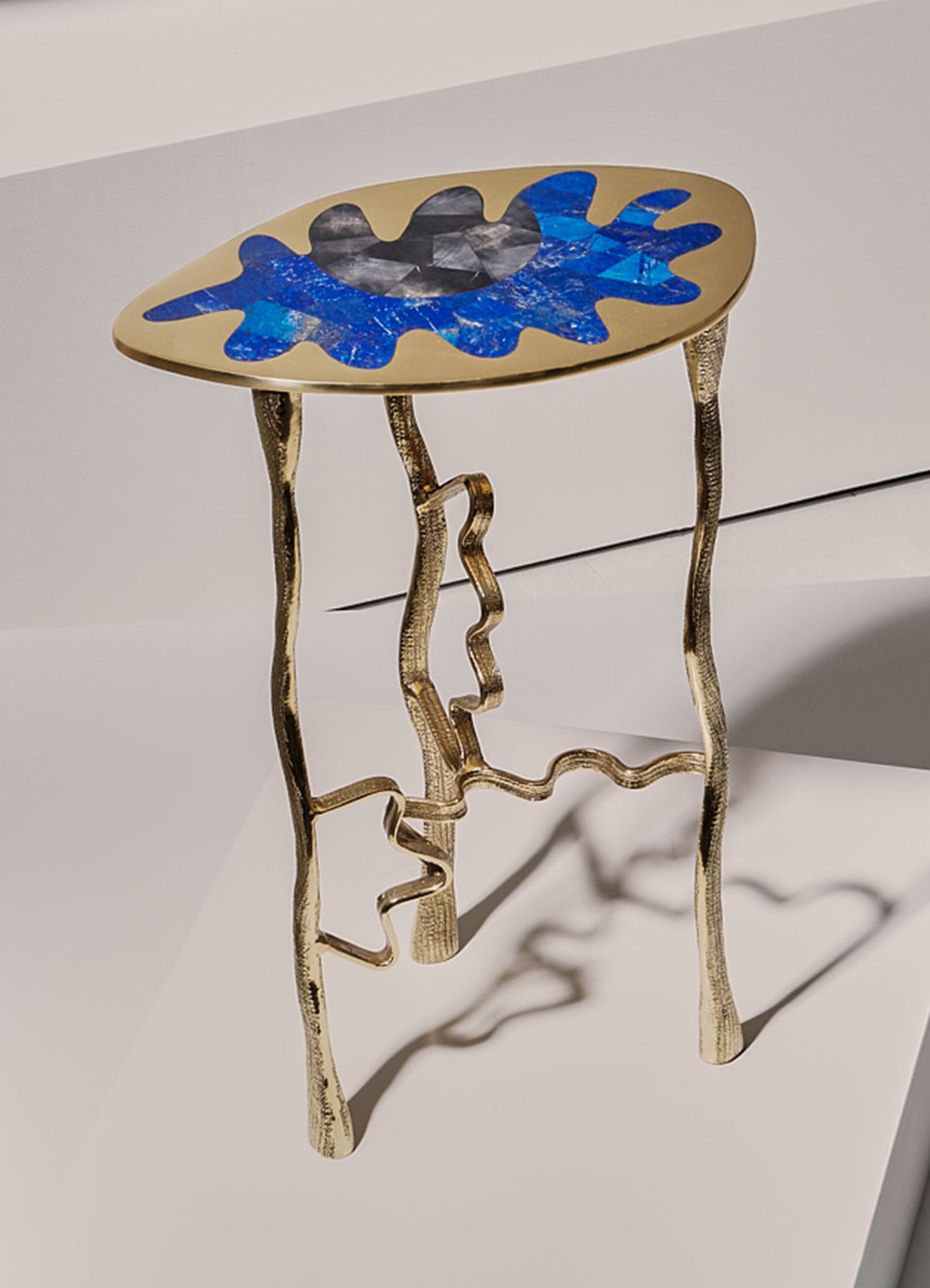 A small side table with a round top featuring a blue and black abstract pattern, supported by an irregular gold metal base with wavy, organic lines. The design combines contemporary style with artistic motifs and metallic finish.