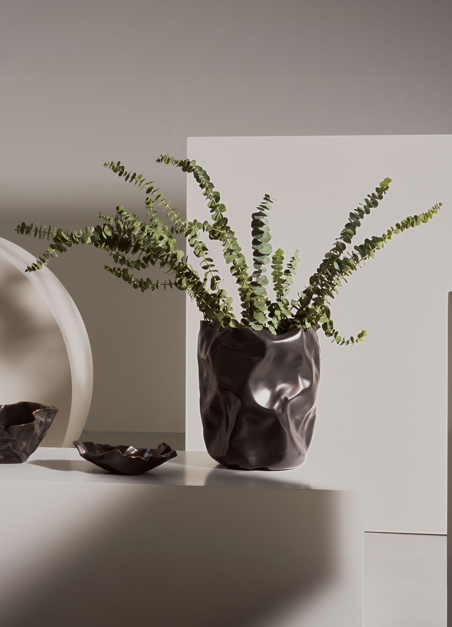A ceramic vase with a textured, irregular surface holding a sprig of green eucalyptus leaves, positioned on a white surface against a minimal background.