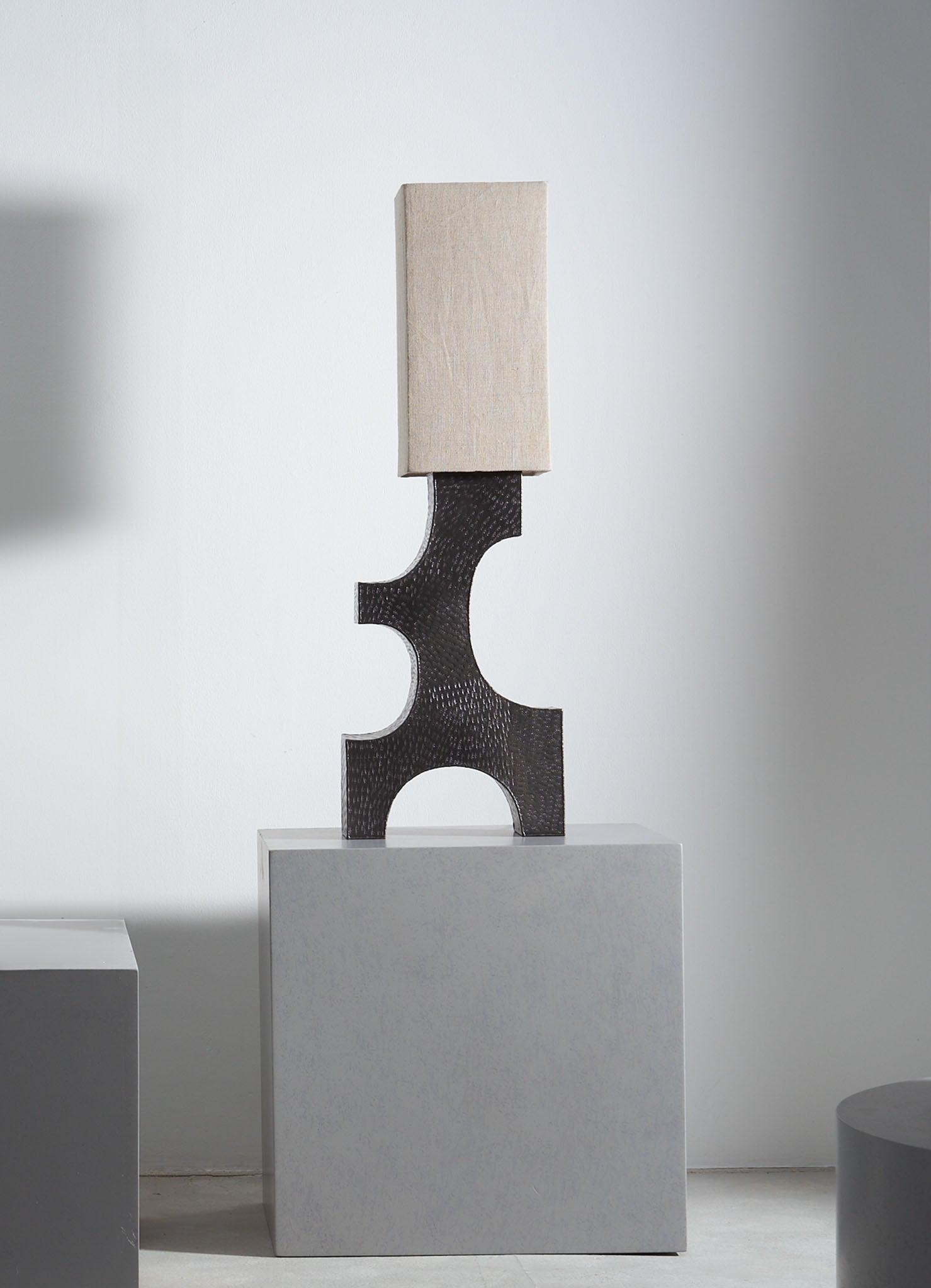 A modern table lamp featuring a black textured base with curved cutouts and a rectangular beige fabric lampshade, positioned on a white pedestal against a plain white background.