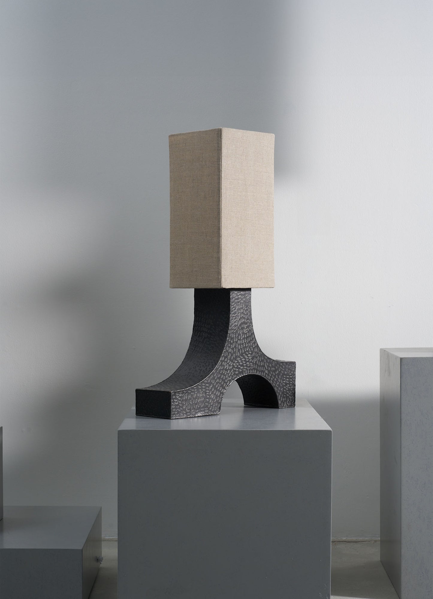A table lamp with a textured black ceramic base and a rectangular beige fabric lampshade, set on a white pedestal against a plain wall.