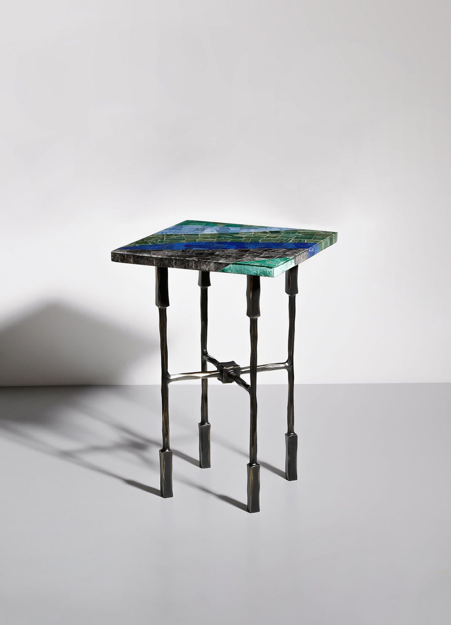 A side table with a rectangular marble top featuring green, blue, and black veining, supported by sculptural, black metal legs with a geometric design and crossbars.