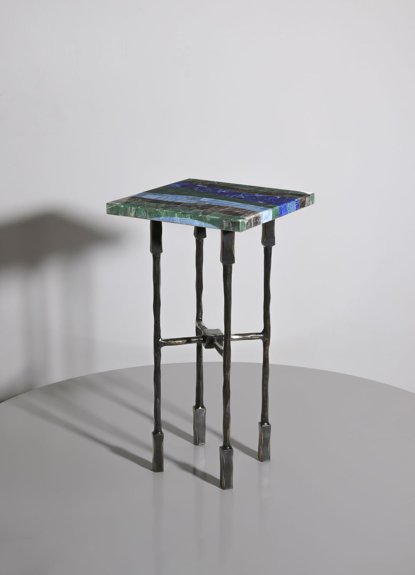 A small side table with a rectangular top made of multicolored marble in shades of green, blue, and black, supported by four black distorted metal legs. The design combines modern and artisanal elements with a focus on natural materials.