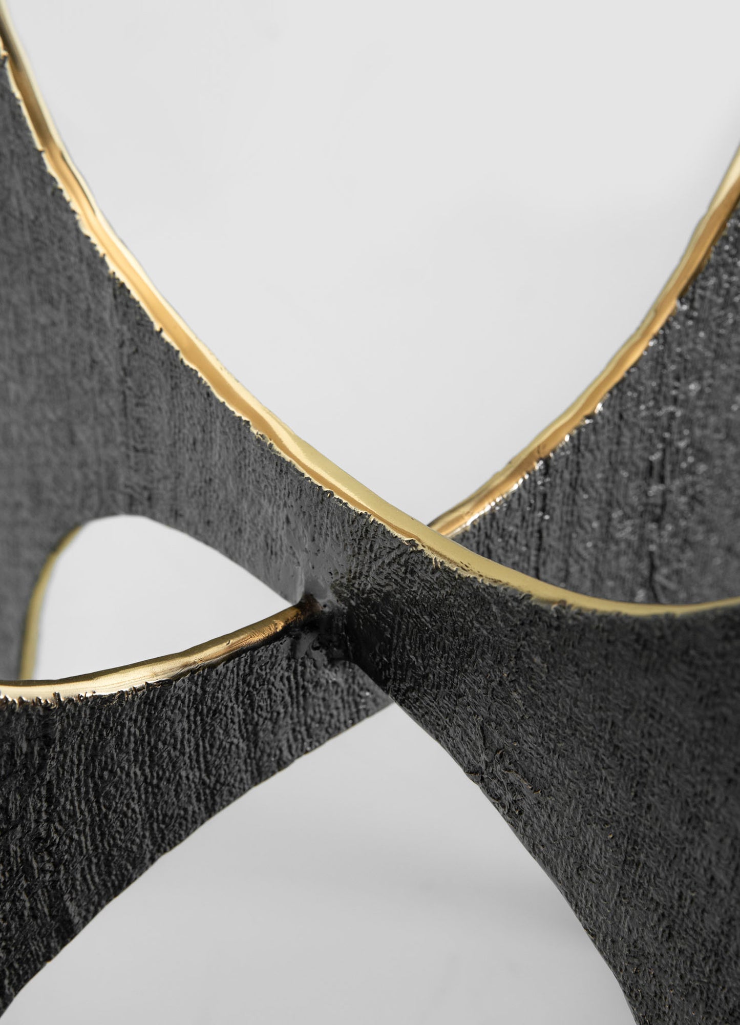 Close-up of a decorative object featuring black textured material with metallic gold accents along the edges, forming an intersecting curved design. The material appears rough and matte, contrasting with the smooth, shiny gold trim.