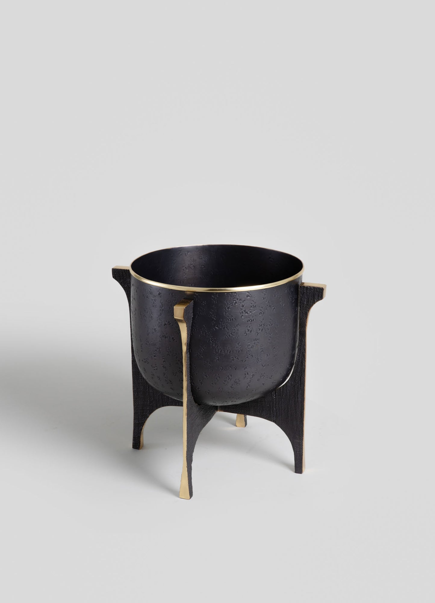 A black metal vessel with a textured matte finish, supported by four curved legs with gold accents, featuring a minimalist design on a plain light background.
