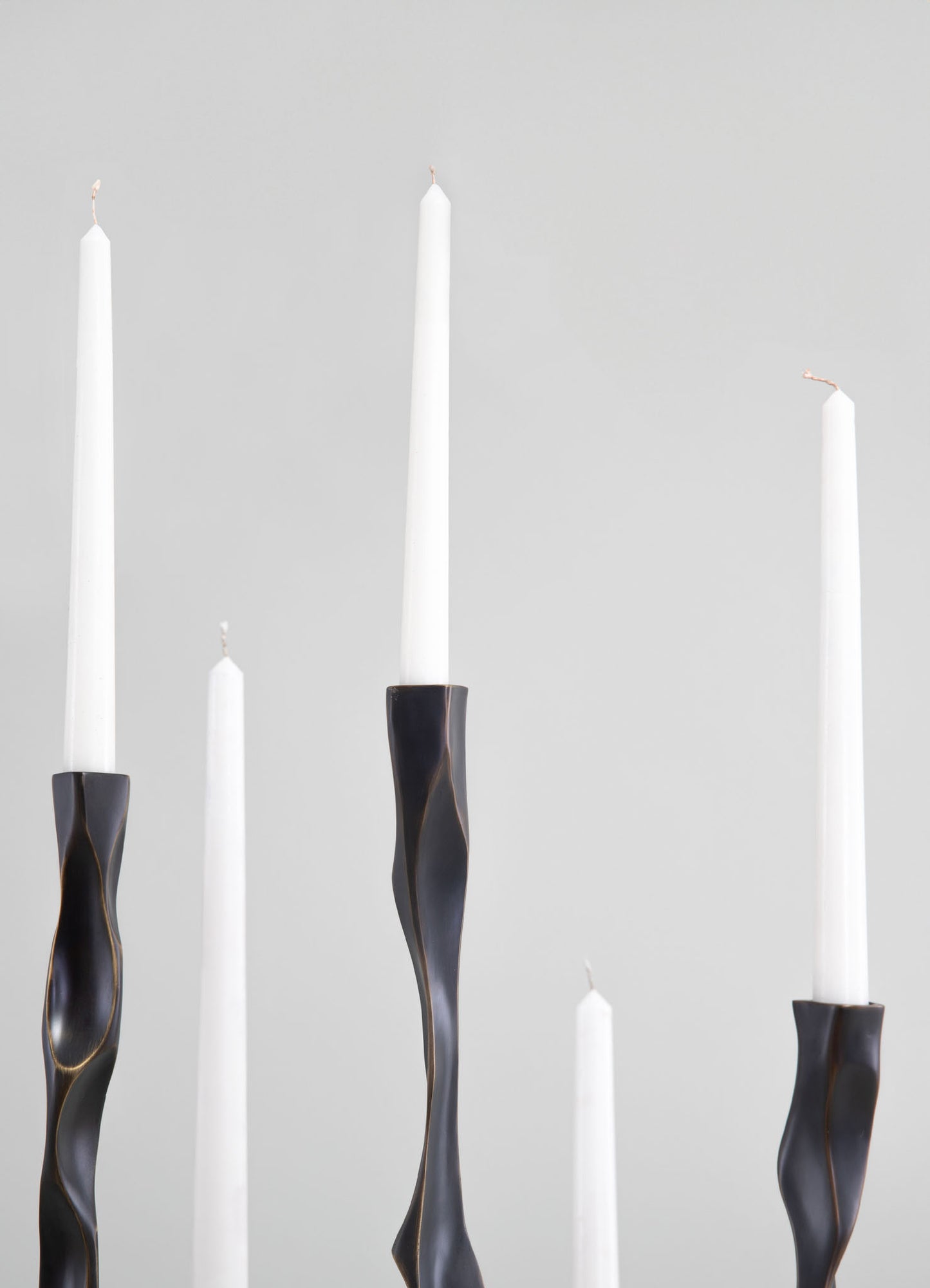 Tall white taper candles with smooth surfaces, held in black sculptural candle holders with twisted, organic shapes, set against a plain light gray background.