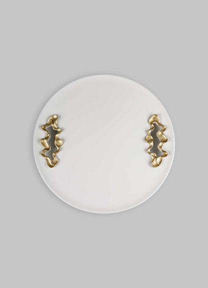 A round white ceramic tray with abstract brass handles featuring organic, irregular shapes, set against a light grey background.