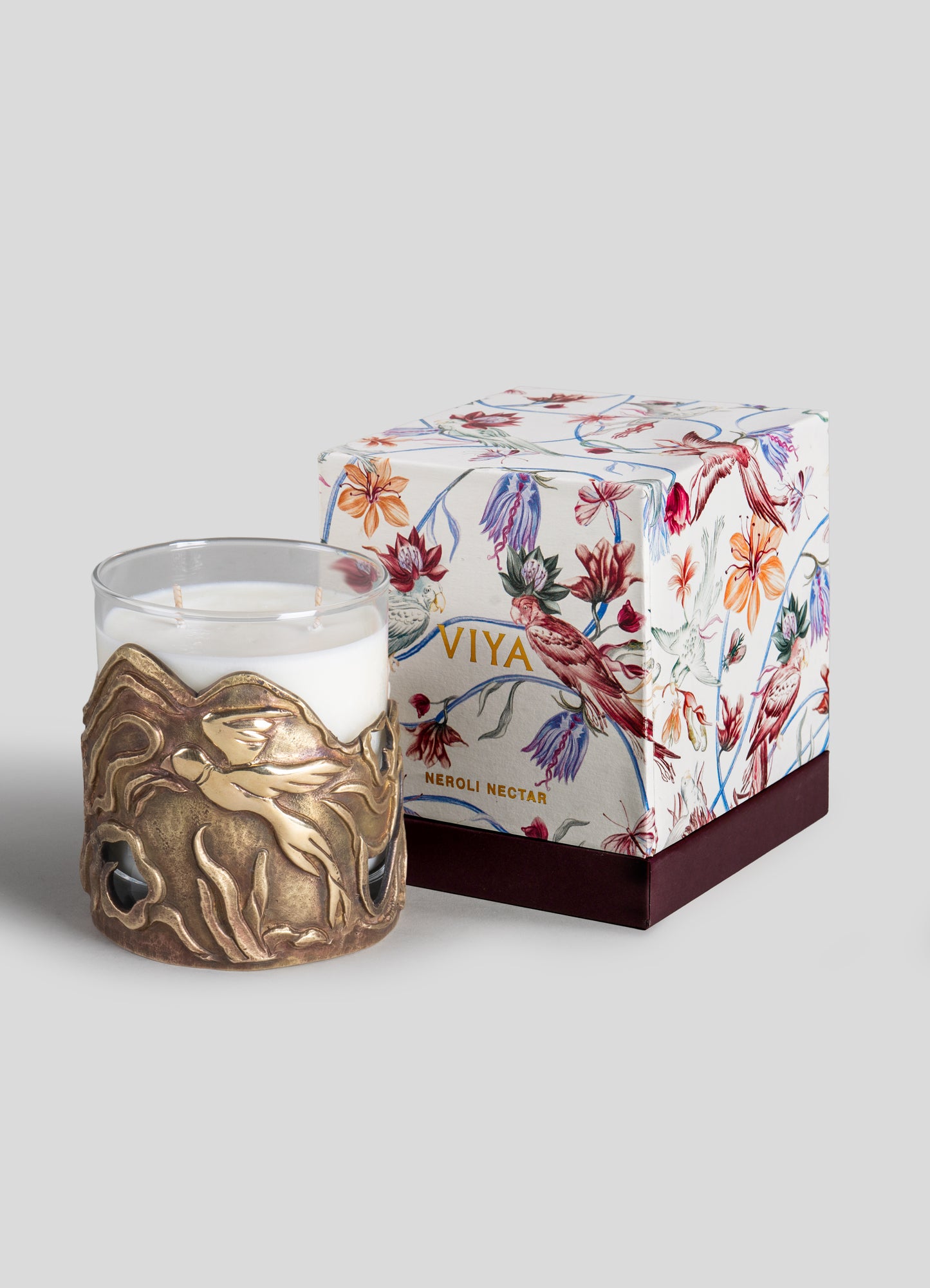 A white scented candle in a glass holder with a brass-like decorative metal sleeve featuring abstract organic shapes. It is paired with a floral-themed gift box with colorful botanical illustrations and the label "Viya Neroli Nectar."