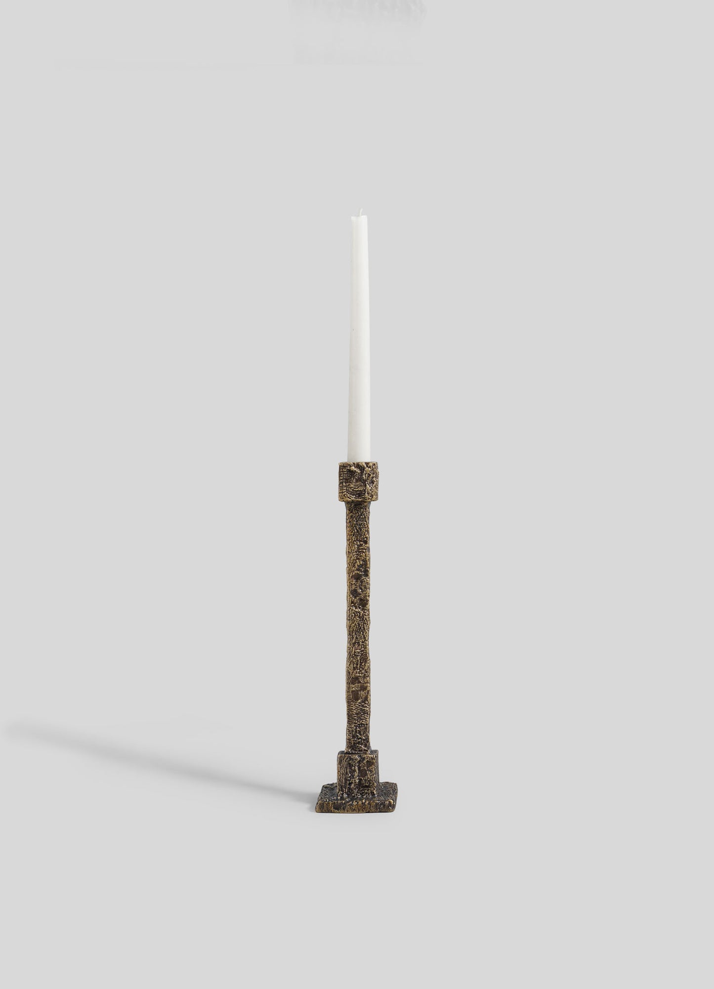 The VIYA Tiruppur Candle Holder Medium, featuring a textured bronze finish and square base inspired by brocade patterns, holds a single white taper candle and is set against a plain light gray background.