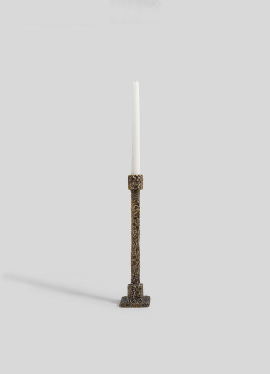 The VIYA Tiruppur Candle Holder Medium, featuring a textured bronze finish and square base inspired by brocade patterns, holds a single white taper candle and is set against a plain light gray background.