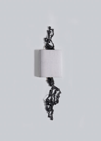 The Andaman Wall Sconce B by VIYA features a cylindrical white form on a light grey background, with irregular black accents—resembling organic shapes—extending from its top and bottom.