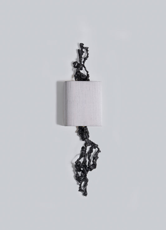 The Andaman Wall Sconce B by VIYA features a cylindrical white form on a light grey background, with irregular black accents—resembling organic shapes—extending from its top and bottom.