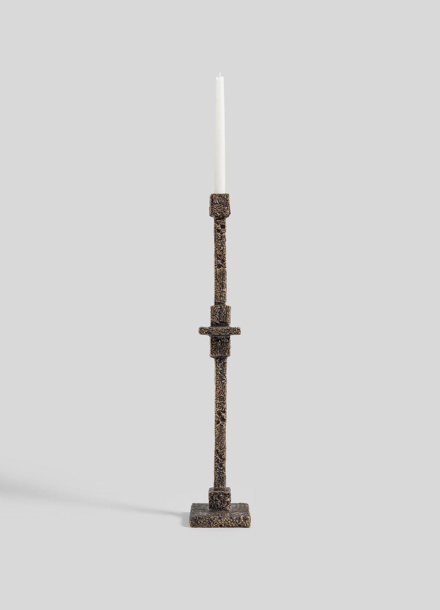 The VIYA Tiruppur Totem Candle Holder Small, a tall textured bronze piece with geometric patterns inspired by Indian brocades, holds a white taper candle against a plain light gray background.