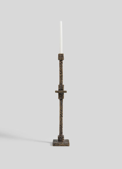 The VIYA Tiruppur Totem Candle Holder Small, a tall textured bronze piece with geometric patterns inspired by Indian brocades, holds a white taper candle against a plain light gray background.