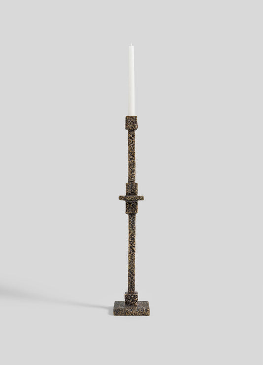 The VIYA Tiruppur Totem Candle Holder Small, a tall textured bronze piece with geometric patterns inspired by Indian brocades, holds a white taper candle against a plain light gray background.