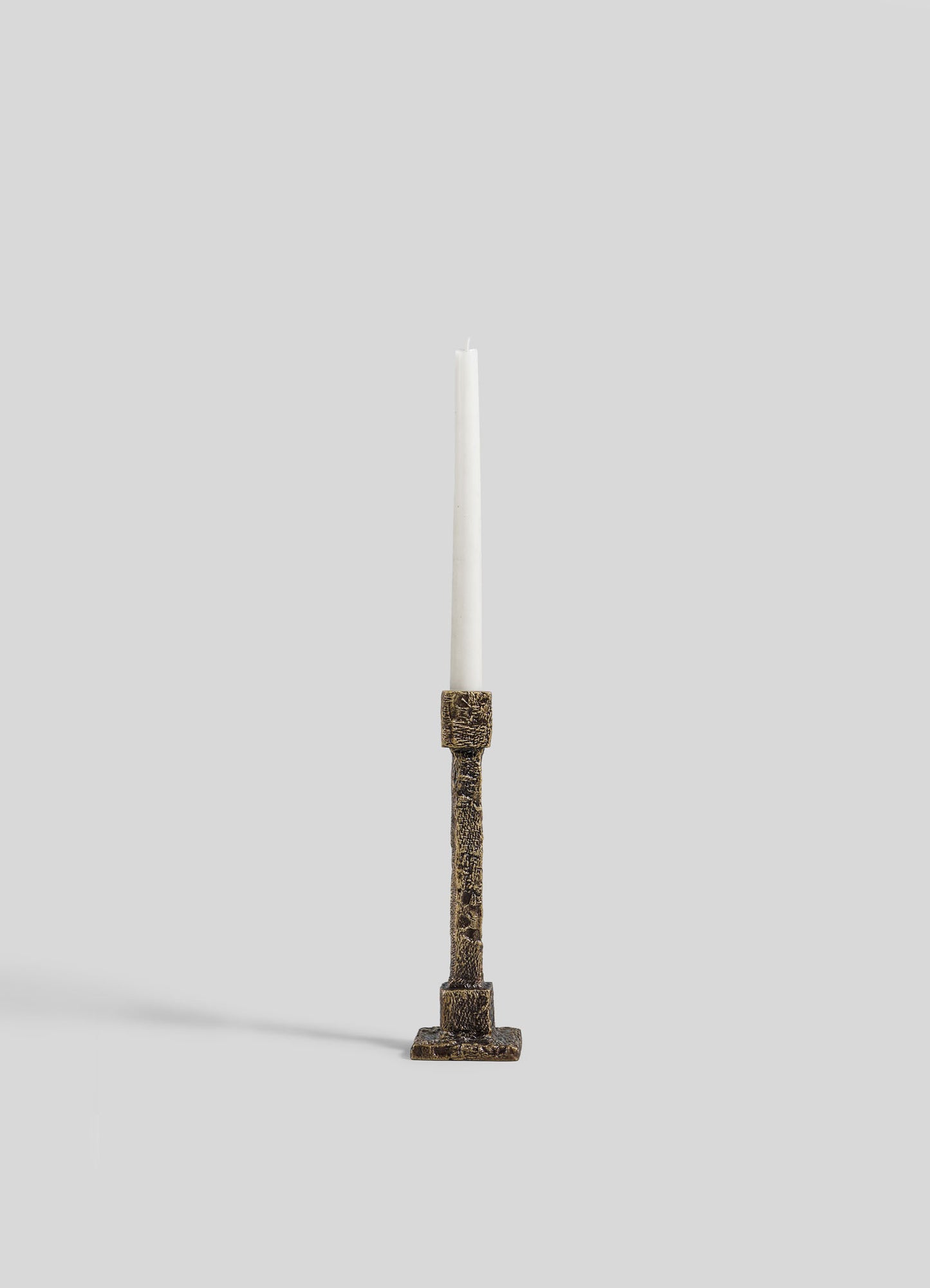 A single white taper candle sits in the VIYA Tiruppur Candle Holder Small, featuring ornate textured bronze inspired by Indian brocades, displayed against a plain light gray background.