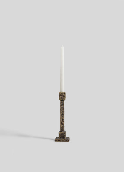 A single white taper candle sits in the VIYA Tiruppur Candle Holder Small, featuring ornate textured bronze inspired by Indian brocades, displayed against a plain light gray background.