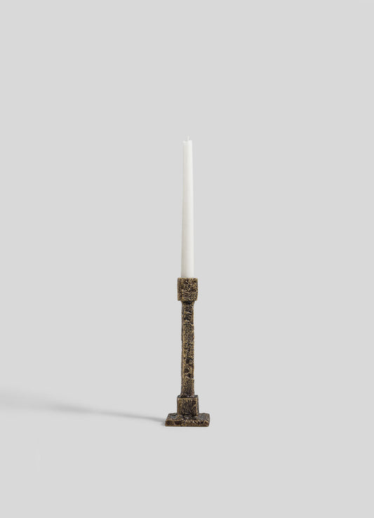 A single white taper candle sits in the VIYA Tiruppur Candle Holder Small, featuring ornate textured bronze inspired by Indian brocades, displayed against a plain light gray background.