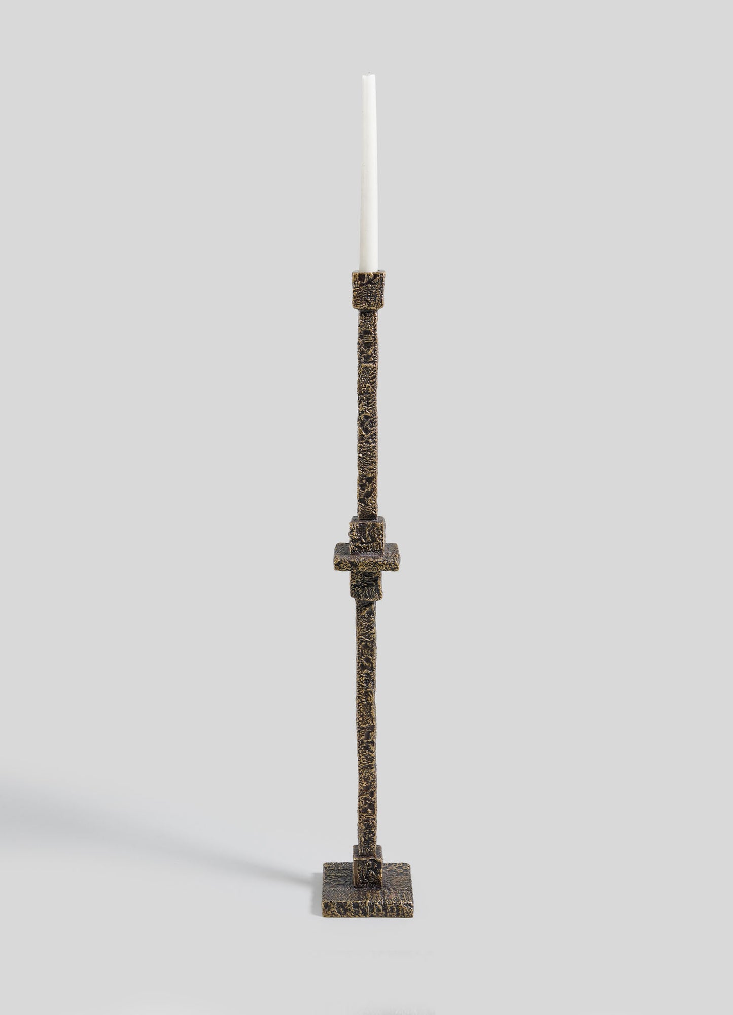 The VIYA Tiruppur Totem Candle Holder Large features a tall, textured bronze design with a square base and slender stem, elegantly displaying a single white taper candle against a plain light gray background.