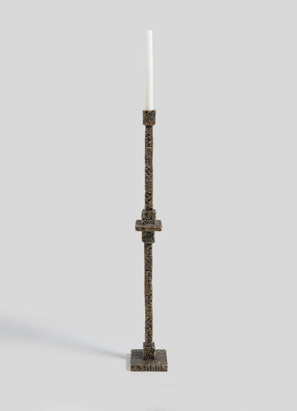 The VIYA Tiruppur Totem Candle Holder Large features a tall, textured bronze design with a square base and slender stem, elegantly displaying a single white taper candle against a plain light gray background.