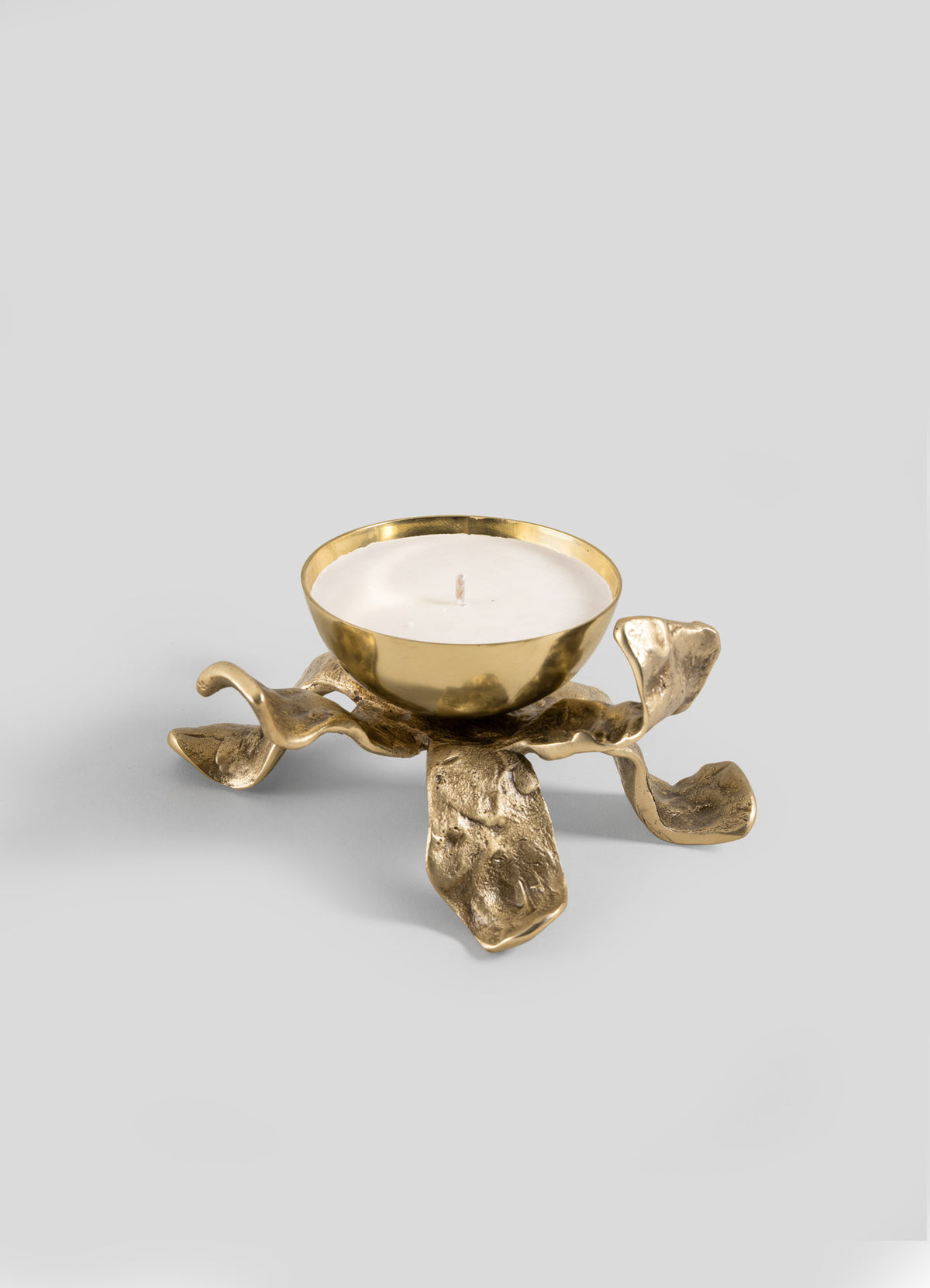 The Viya Glory Lily Scented Diya features a gold metal holder with a bowl-shaped top cradling a white tealight candle. Its sculptural base, designed with four organic, wavy, petal-like legs, creates an artistic modern heirloom look set against a plain gray background.