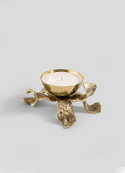 The Viya Glory Lily Scented Diya features a gold metal holder with a bowl-shaped top cradling a white tealight candle. Its sculptural base, designed with four organic, wavy, petal-like legs, creates an artistic modern heirloom look set against a plain gray background.