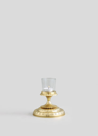 A gold-colored metal candle holder with a fluted base and a scalloped edge top, featuring a transparent glass cup with a lit white tea light candle. The design is elegant and classical, with a polished finish.