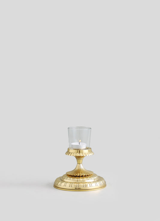 A gold-colored metal candle holder with a fluted base and a scalloped edge top, featuring a transparent glass cup with a lit white tea light candle. The design is elegant and classical, with a polished finish.