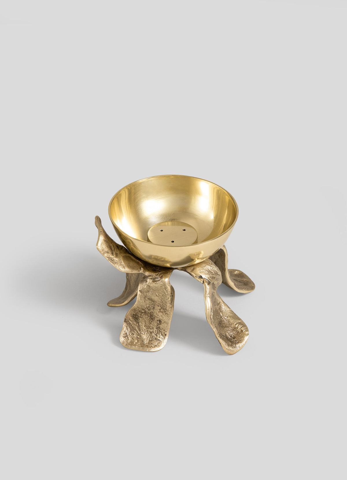 The Glory Lily Incense Diya by VIYA is a gold-colored metal bowl featuring a textured, organic-shaped base reminiscent of petals or leaves—expertly crafted using ancient metalworking techniques—and presented against a plain light gray background.