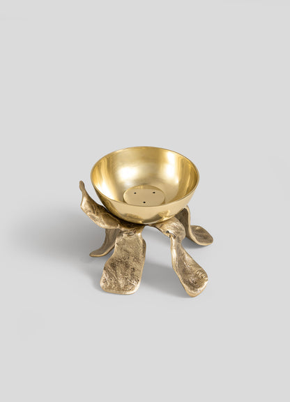 The Glory Lily Incense Diya by VIYA is a gold-colored metal bowl featuring a textured, organic-shaped base reminiscent of petals or leaves—expertly crafted using ancient metalworking techniques—and presented against a plain light gray background.