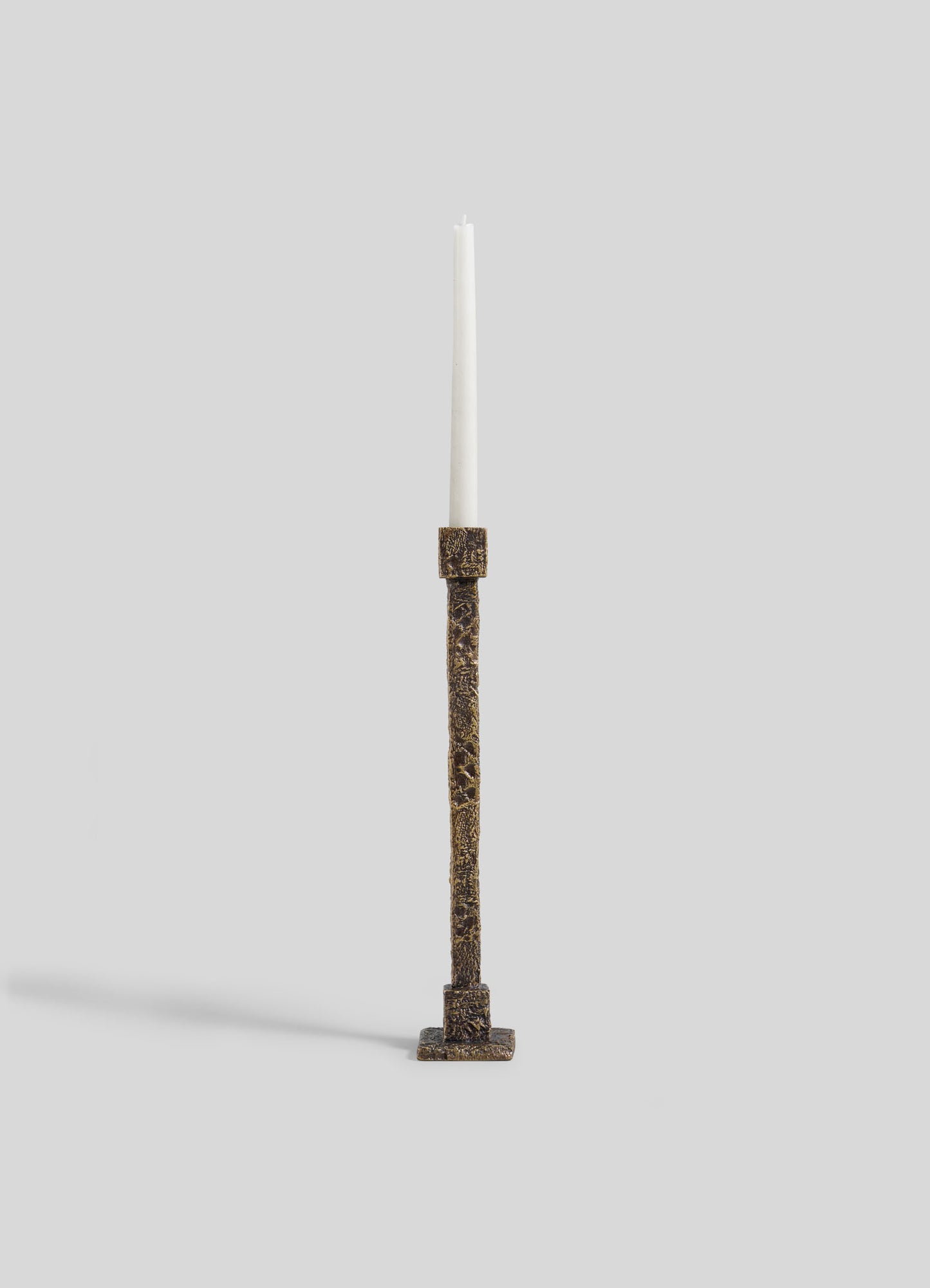 A tall white candle is displayed in the VIYA Tiruppur Candle Holder Large, a textured, rectangular bronze piece inspired by brocades and handwoven fabrics, set against a plain light gray background.