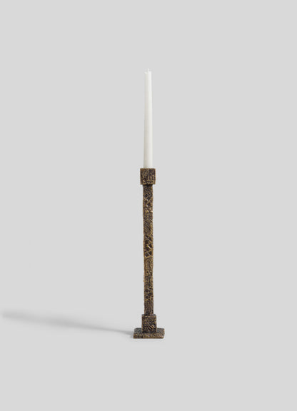 A tall white candle is displayed in the VIYA Tiruppur Candle Holder Large, a textured, rectangular bronze piece inspired by brocades and handwoven fabrics, set against a plain light gray background.