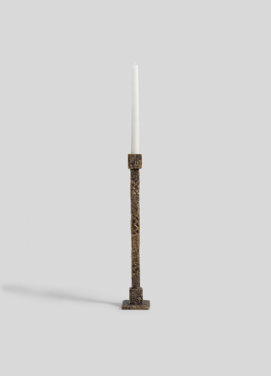 A tall white candle is displayed in the VIYA Tiruppur Candle Holder Large, a textured, rectangular bronze piece inspired by brocades and handwoven fabrics, set against a plain light gray background.