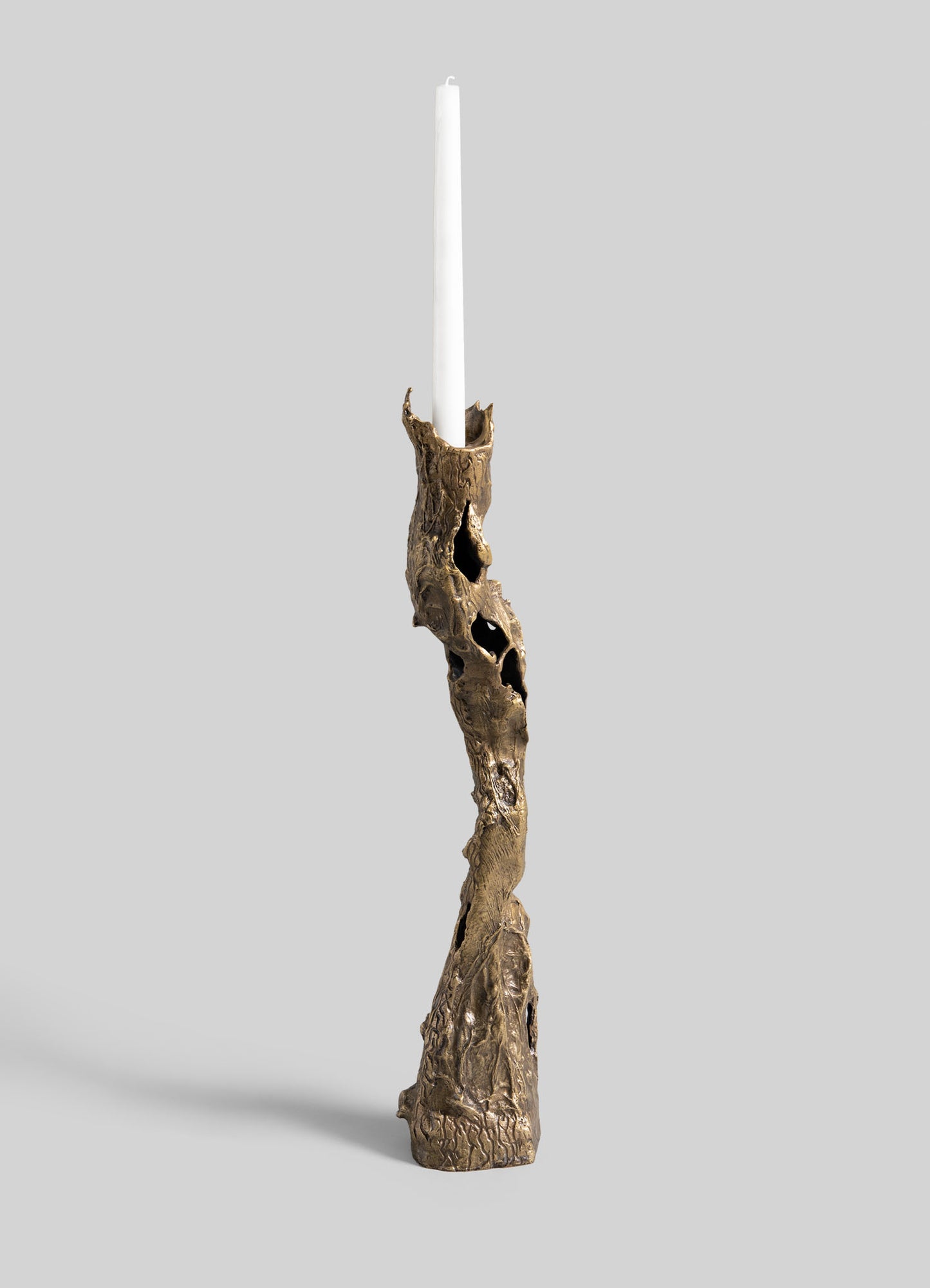 A tall, white candle sits in the VIYA Kikar Candle Holder Large—a sculptural, twisted brass piece with a textured, hollowed design—set against a plain light gray background.