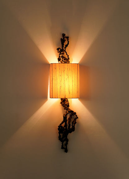 The Andaman Wall Sconce B by VIYA features a rectangular beige shade that emits a warm glow, with decorative dark metal accents inspired by brass objects above and below, casting patterned shadows on the wall.
