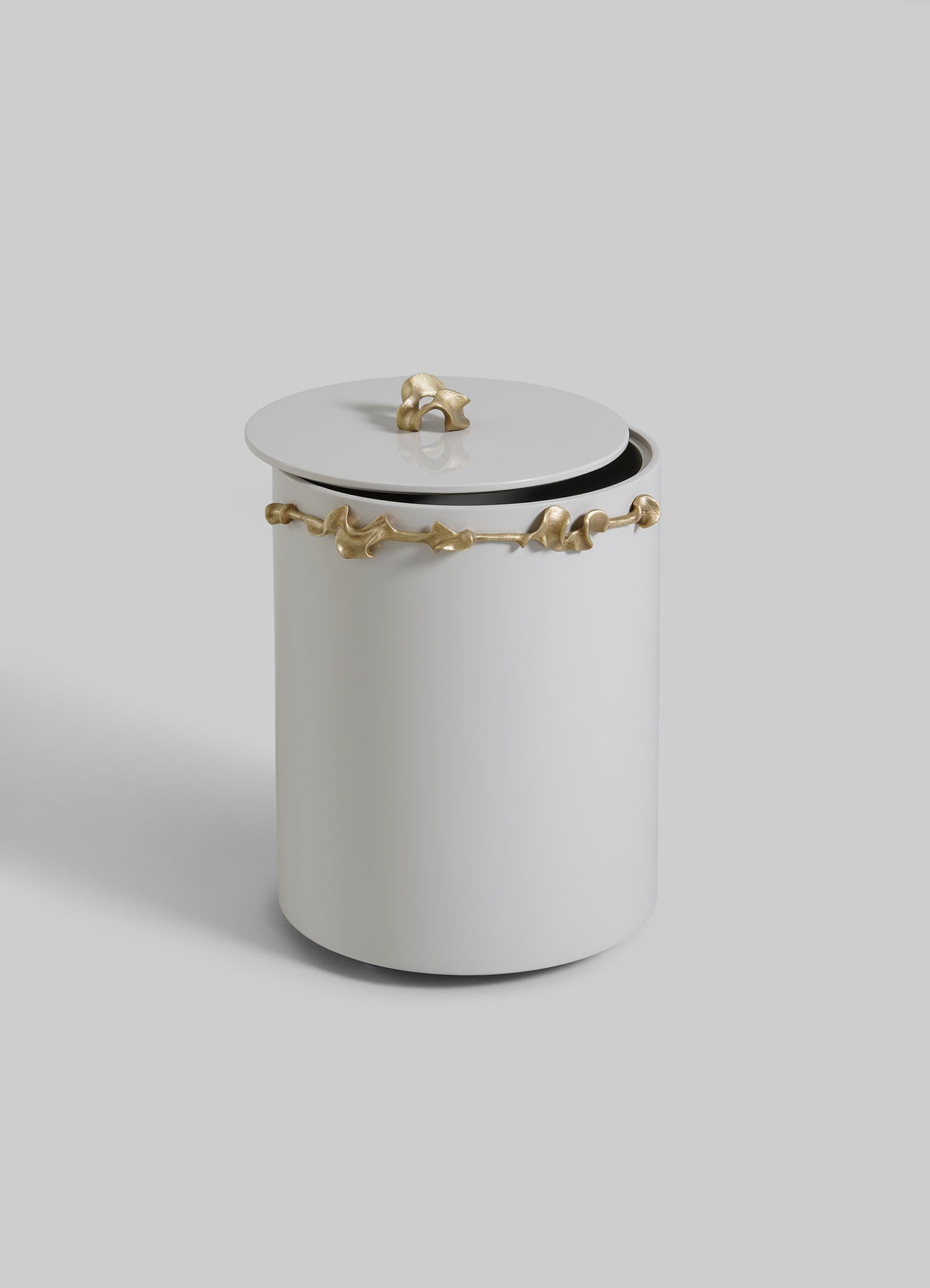 The VIYA Saffron Ecru Bin With Lid is a white cylindrical container with gold accents and vine-like gold details, evoking bronzed decor. The lid sits slightly ajar against a light gray background.