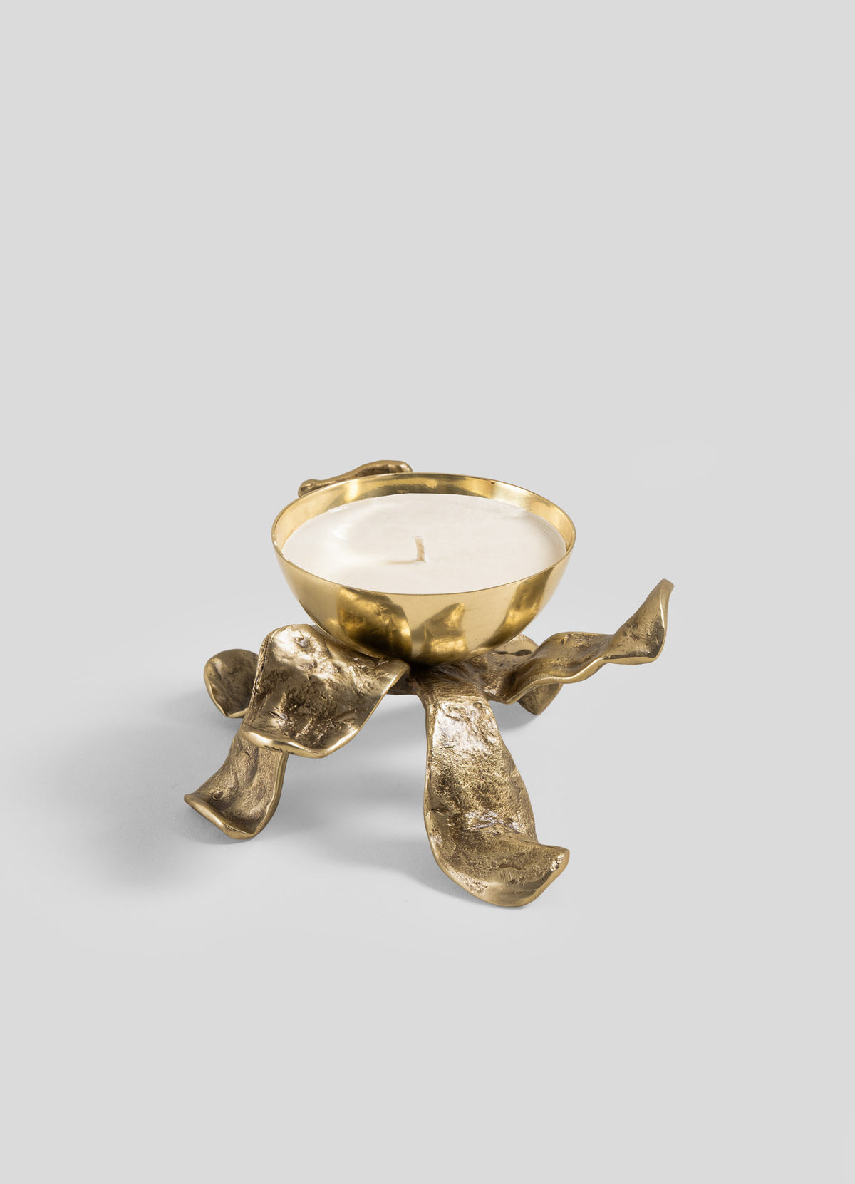 The Viya Glory Lily Scented Diya is a sculptural gold candle holder with a white candle, featuring a bowl-shaped top and four textured, ribbon-like legs—crafted using ancient metalworking techniques—set against a plain light gray background.