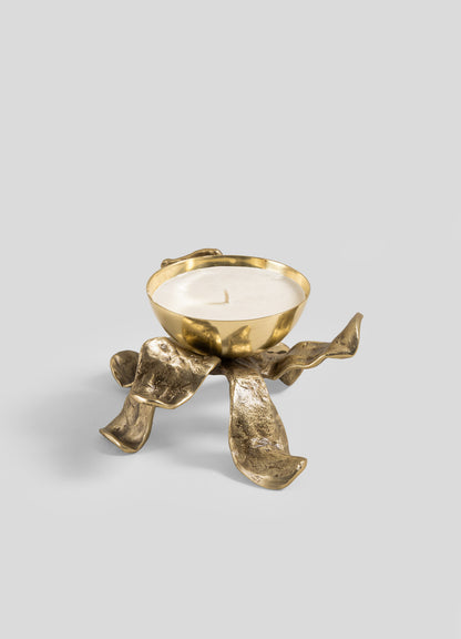 The Viya Glory Lily Scented Diya is a sculptural gold candle holder with a white candle, featuring a bowl-shaped top and four textured, ribbon-like legs—crafted using ancient metalworking techniques—set against a plain light gray background.