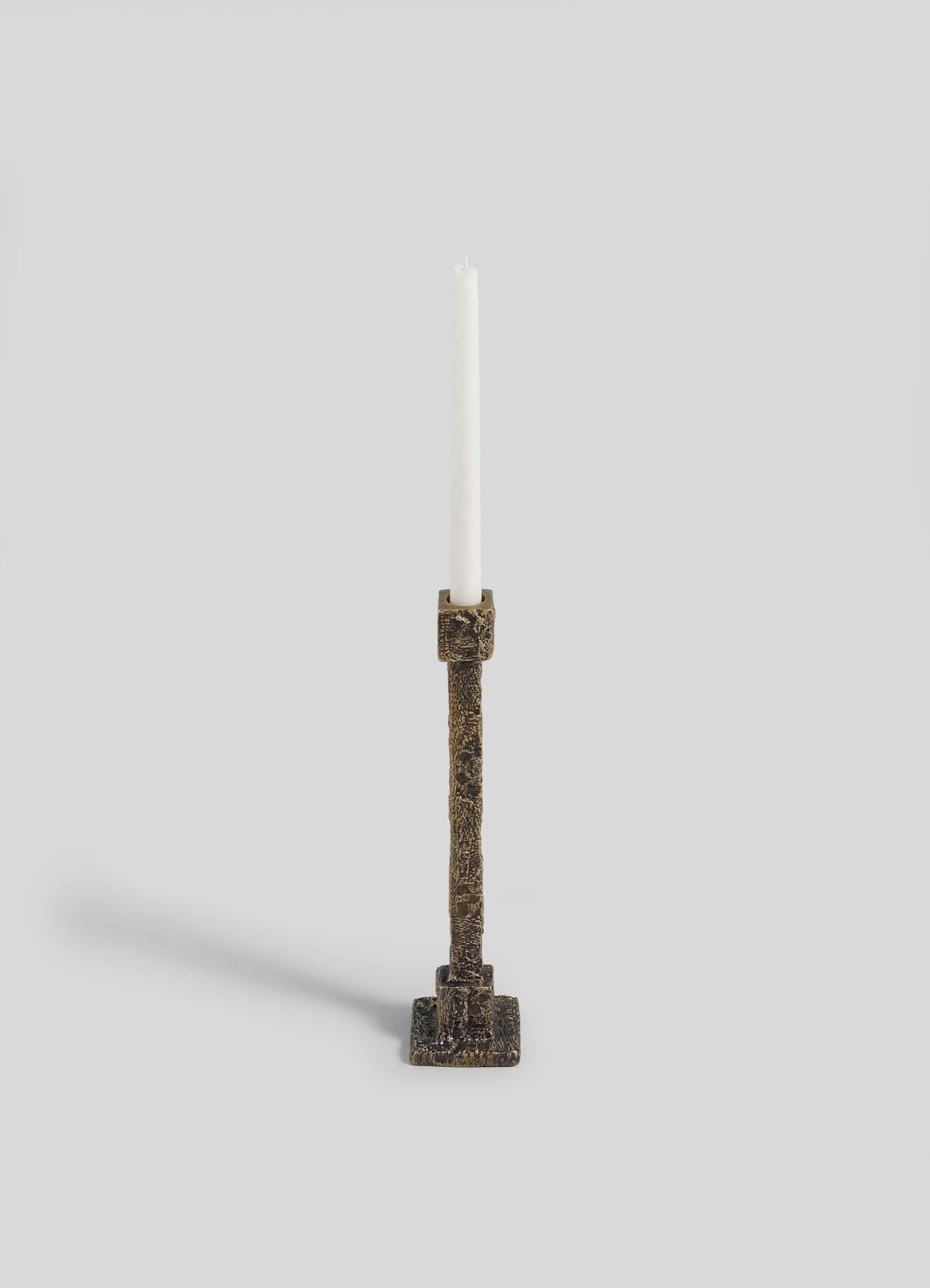 The VIYA Tiruppur Candle Holder Medium, a tall textured bronze piece with a white taper candle and square base, stands elegantly against a light gray background.