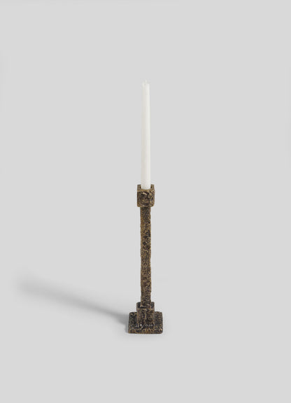 The VIYA Tiruppur Candle Holder Medium, a tall textured bronze piece with a white taper candle and square base, stands elegantly against a light gray background.