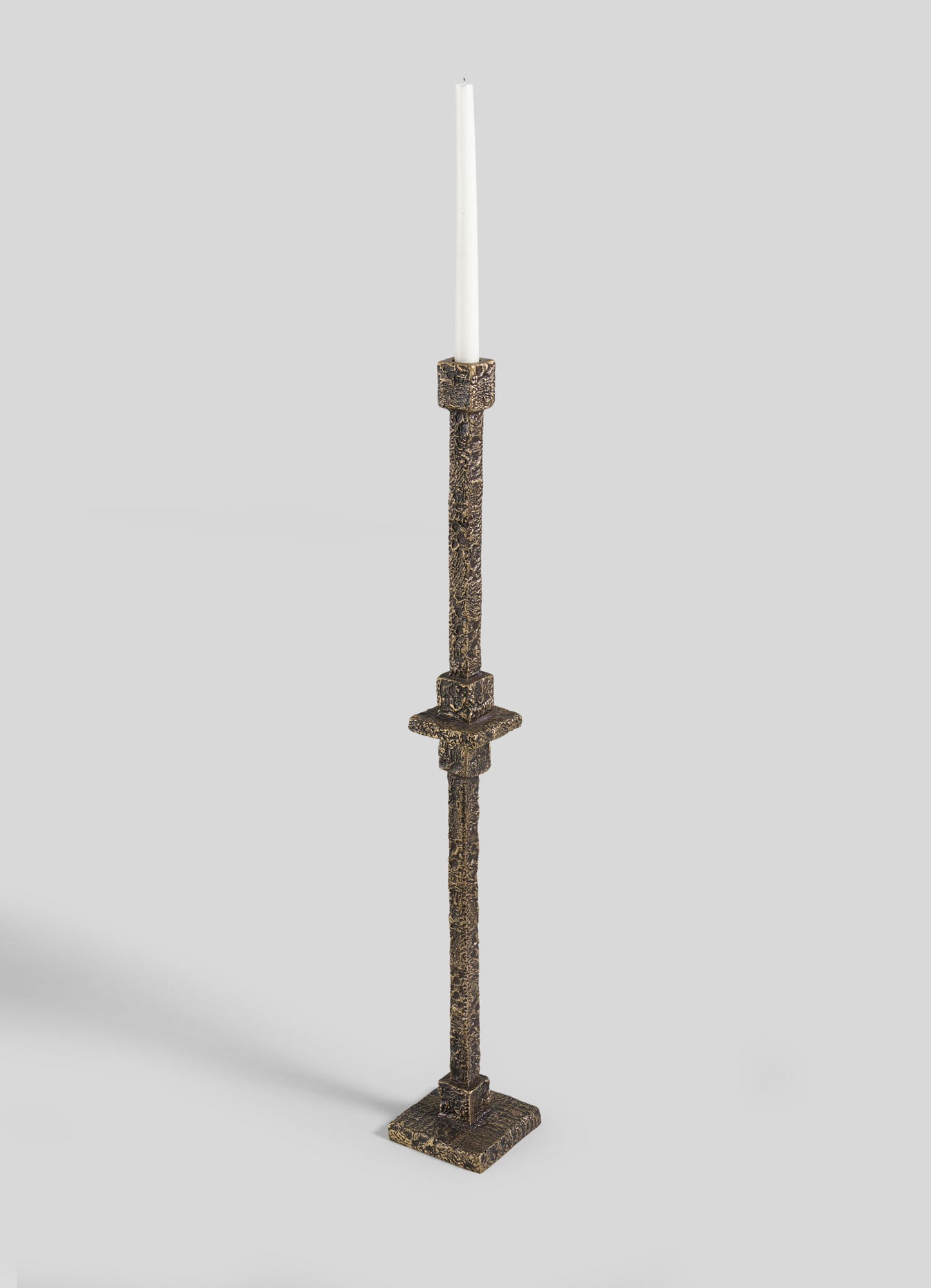The VIYA Tiruppur Totem Candle Holder Large, a tall textured bronze piece with a square base and single white taper candle, evokes handwoven elegance against a plain light gray background.