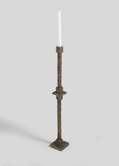 The VIYA Tiruppur Totem Candle Holder Large, a tall textured bronze piece with a square base and single white taper candle, evokes handwoven elegance against a plain light gray background.