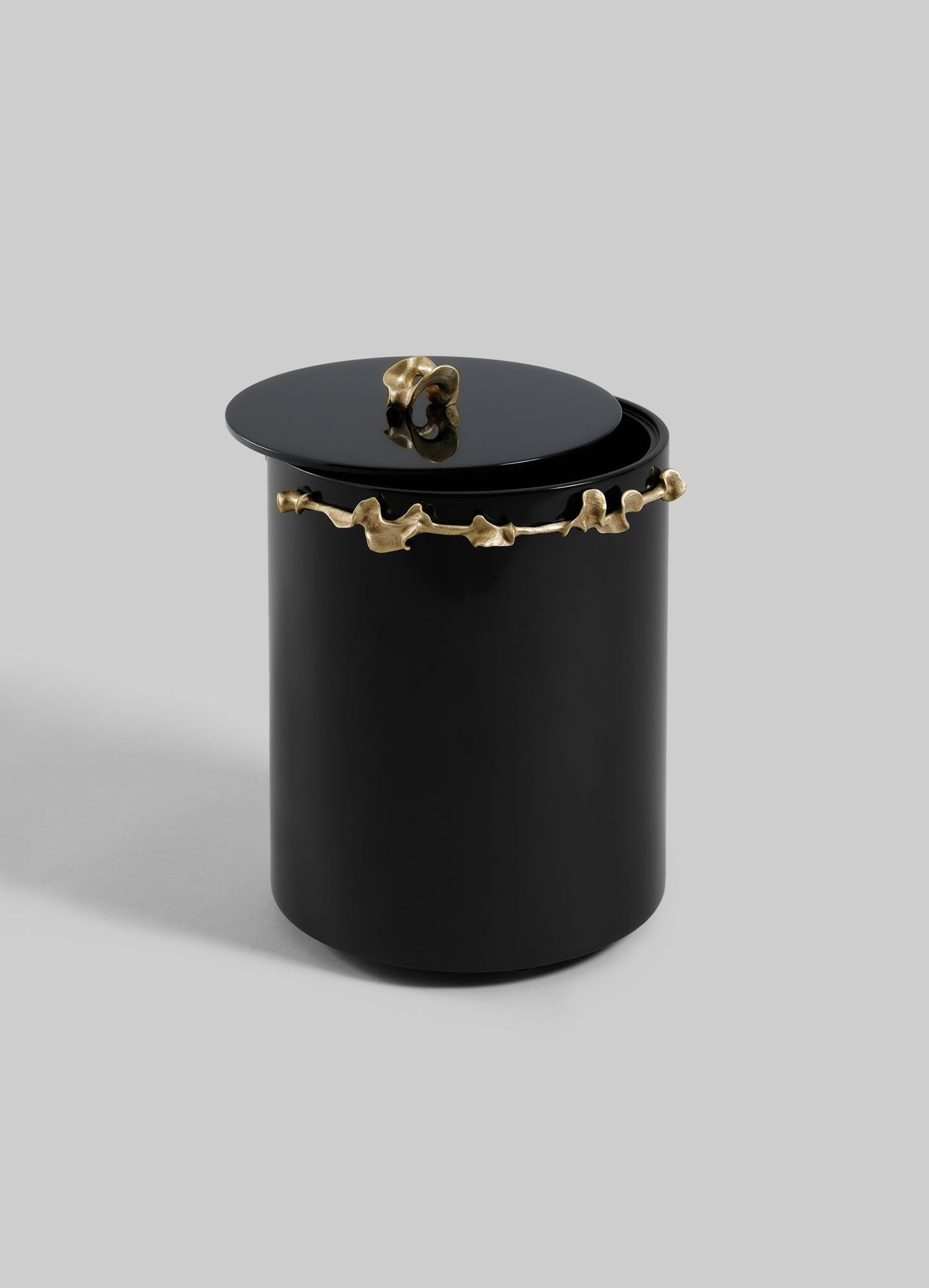 The VIYA Saffron Black Bin With Lid is a matte black cylindrical container with a lid, accented by irregular gold detailing along the upper edge and on the handle, adding contemporary elegance against a plain gray backdrop.
