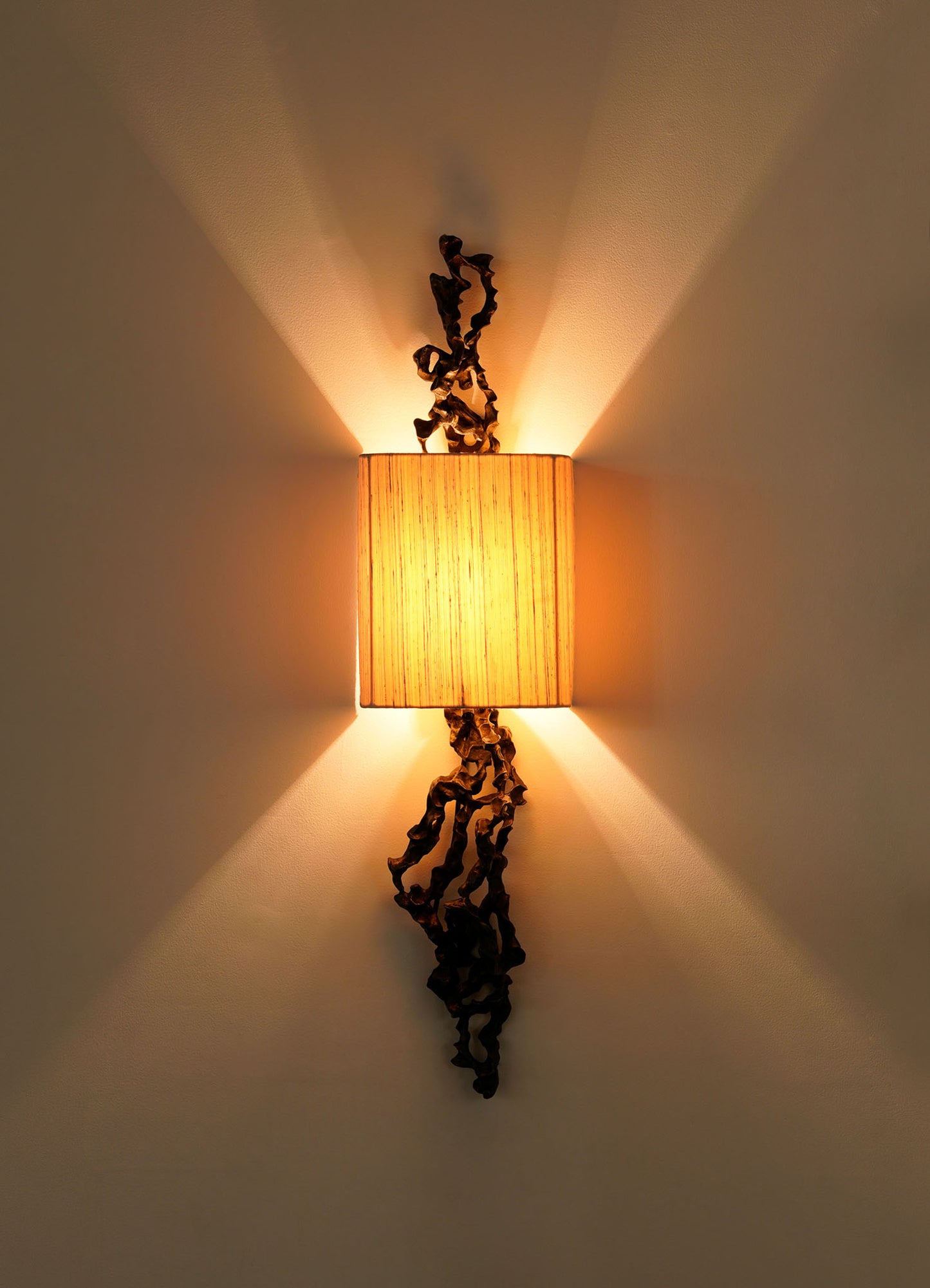 The VIYA Andaman Wall Sconce A features a rectangular beige shade with decorative black metal accents, emitting warm light and casting symmetrical, organic-inspired shadows reminiscent of the Andaman Islands.