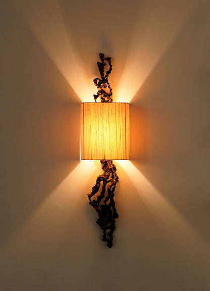 The VIYA Andaman Wall Sconce A features a rectangular beige shade with decorative black metal accents, emitting warm light and casting symmetrical, organic-inspired shadows reminiscent of the Andaman Islands.