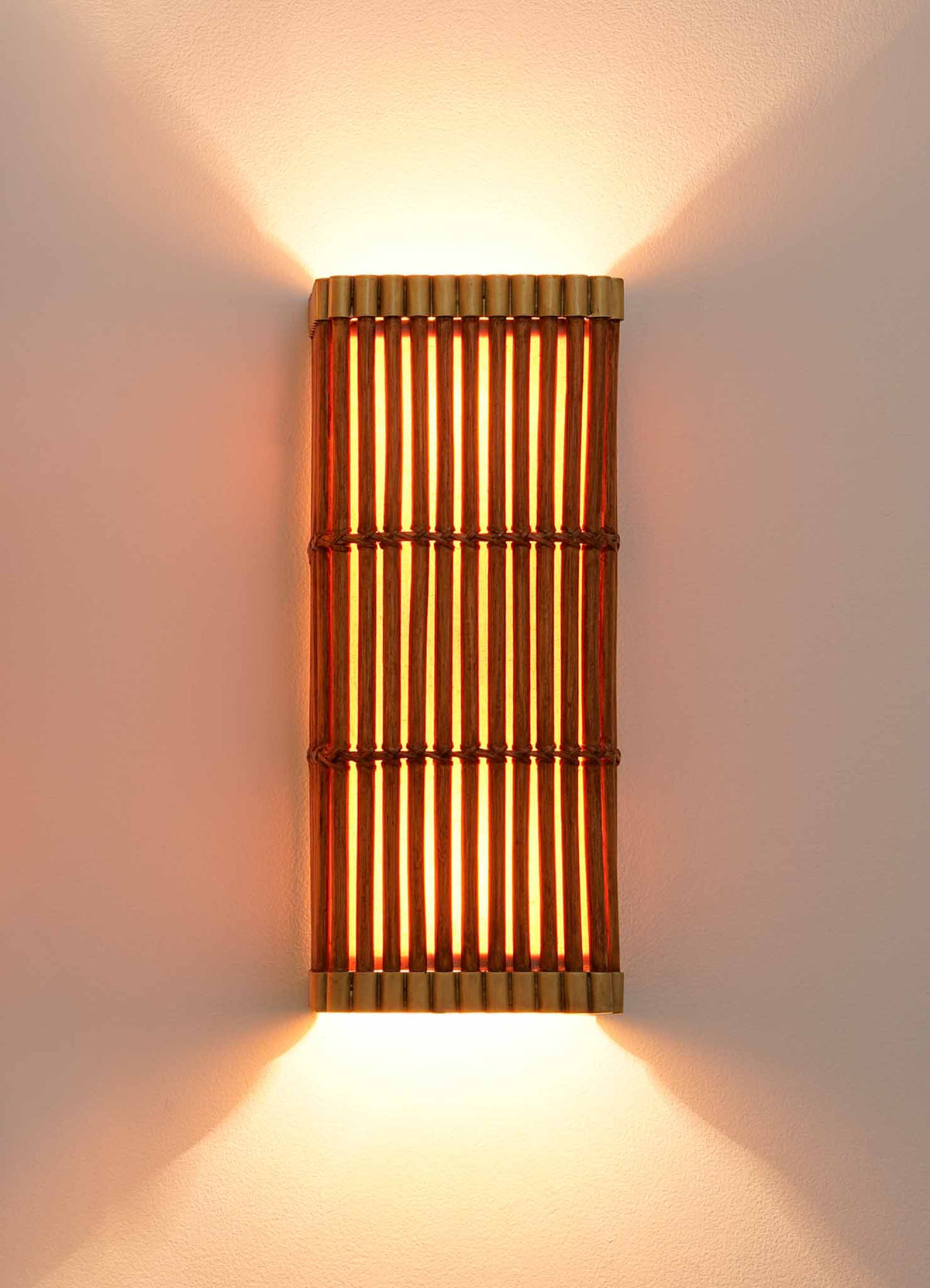 The Namsai Wall Sconce by VIYA, handcrafted using traditional Indian techniques, is a wall-mounted bamboo lighting fixture that emits warm light both upward and downward, softly illuminating the white wall. Featuring vertical wooden slats and gold accents on the top and bottom edges, this sconce creates an inviting glow.