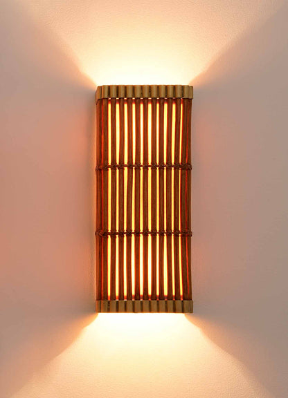 The Namsai Wall Sconce by VIYA, handcrafted using traditional Indian techniques, is a wall-mounted bamboo lighting fixture that emits warm light both upward and downward, softly illuminating the white wall. Featuring vertical wooden slats and gold accents on the top and bottom edges, this sconce creates an inviting glow.