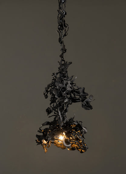 The Gardens Of India Pendant Light by VIYA features dark, intricately twisted metal with organic floral shapes, evoking an Indian garden. A warm bulb glows at its center, set against a plain dark background.